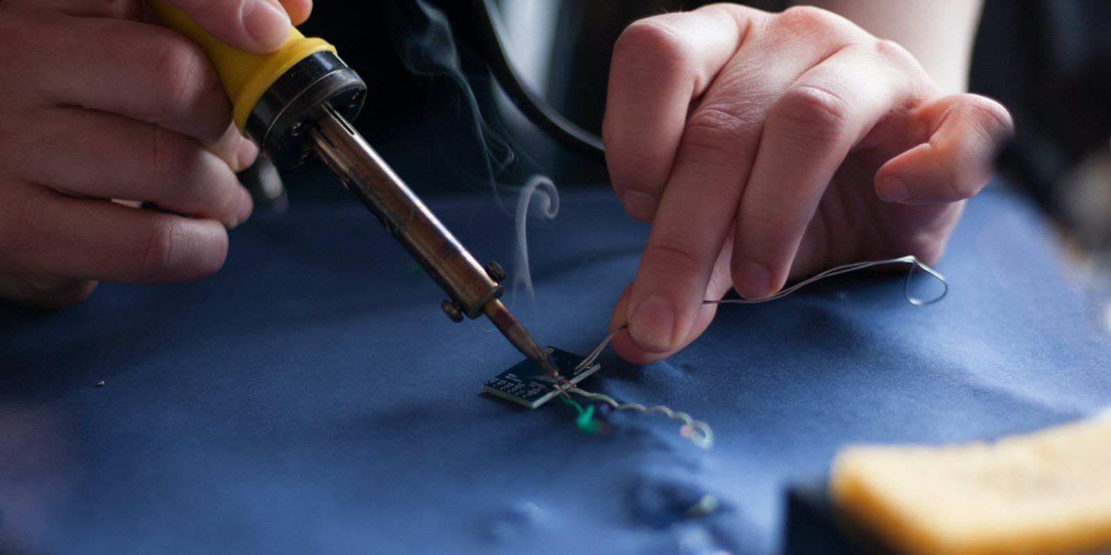 Hands soldering a thin wire to a tiny board on a blue mat.