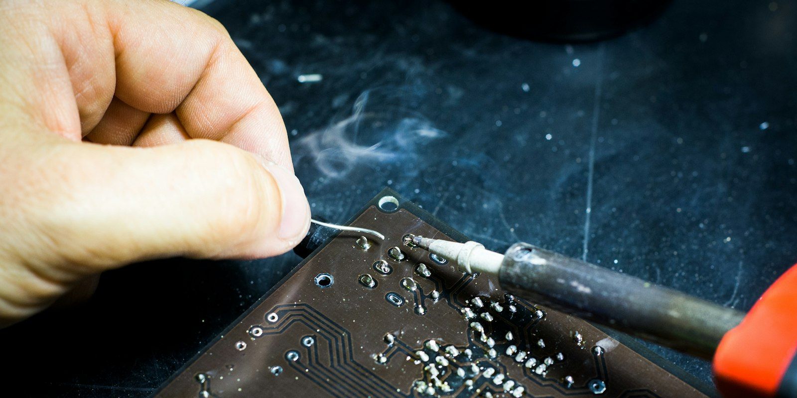 A hand feeding solder to an iron on the underside of a circuit board.