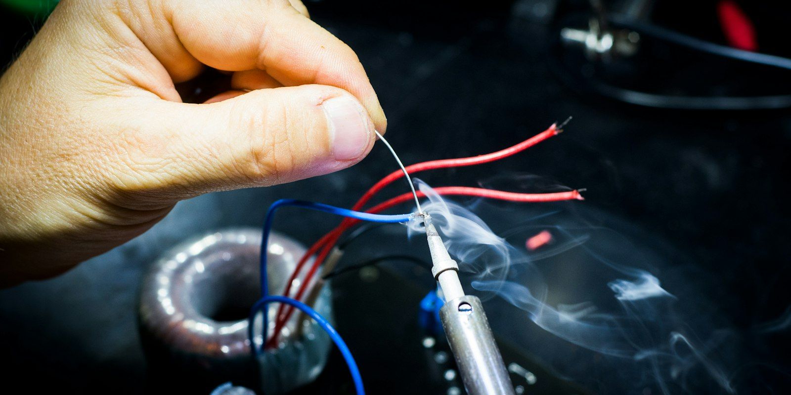 A hand holding a wire against a soldering iron tip, smoke rising over a circuit board.