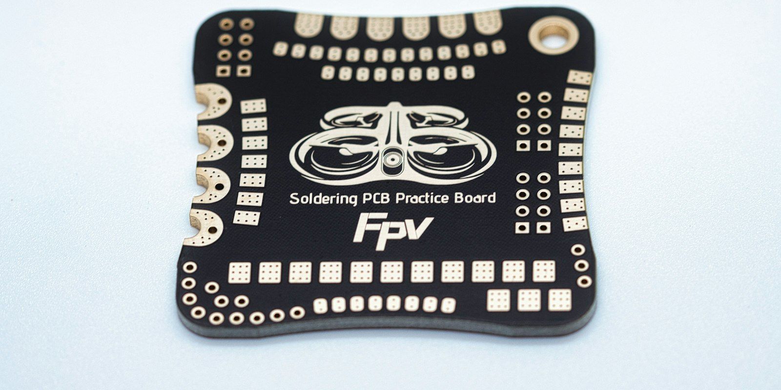 A black soldering practice board with rows of gold pads on a white surface.